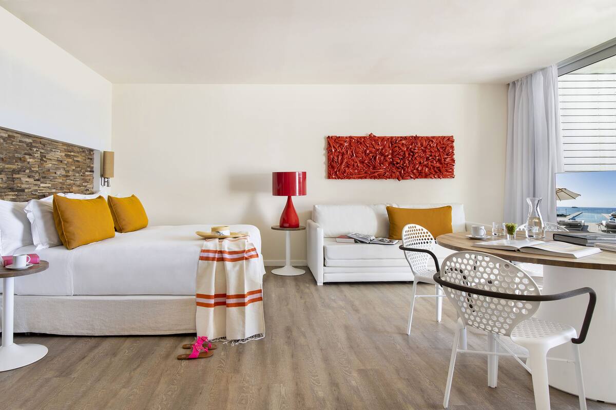 A modern hotel room with a bed, a sofa, a dining table, and a chair. There is a red lamp, a white wall with a textured art piece, and a window with a view.