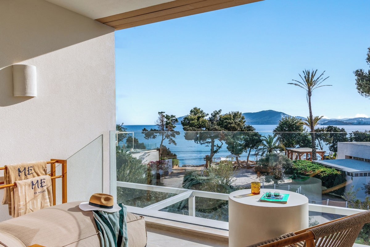 A balcony with a view of the sea, a hat, a towel, and a small table with a book and glasses.
