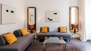 A modern living room with two grey sofas, yellow cushions, a glass coffee table, and wall-mounted mirrors with lighting. - Suite Prado Hotel (Madrid)