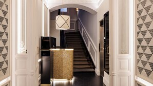 A grand hallway with a staircase, a decorative light fixture, and a reception area with a tiled wall. - Suite Prado Hotel (Madrid)