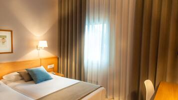 A hotel room with two beds, a bedside table with a lamp, a chair, and a window with curtains.