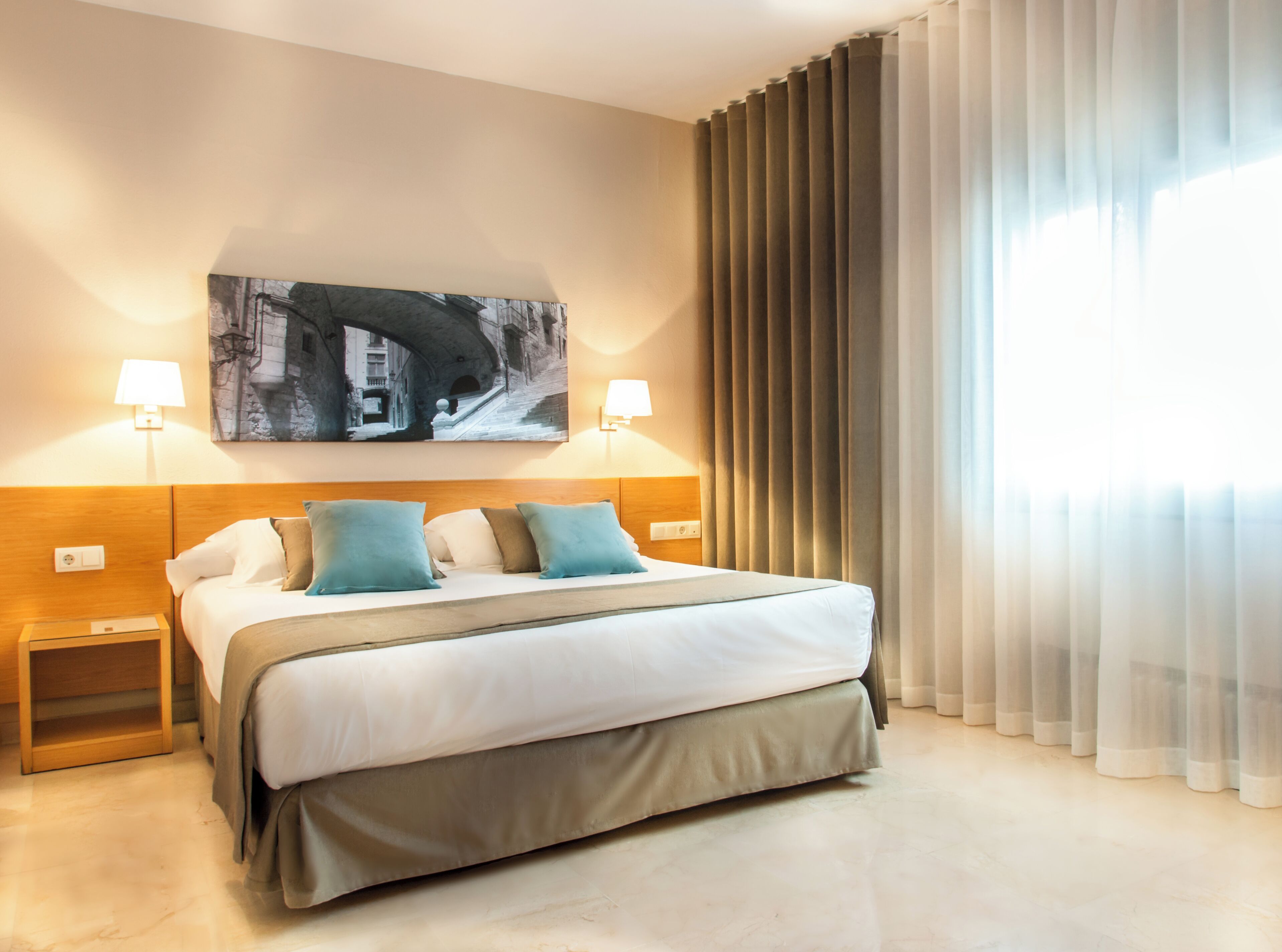 A hotel room with a bed, two bedside lamps, a painting on the wall, and a window with curtains.