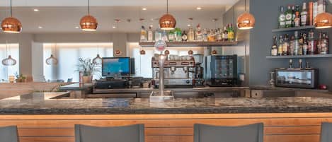 Zona de bar con encimera de granito, taburetes, lámparas colgantes de cobre, máquina de café y estantes con diferentes botellas de licores.