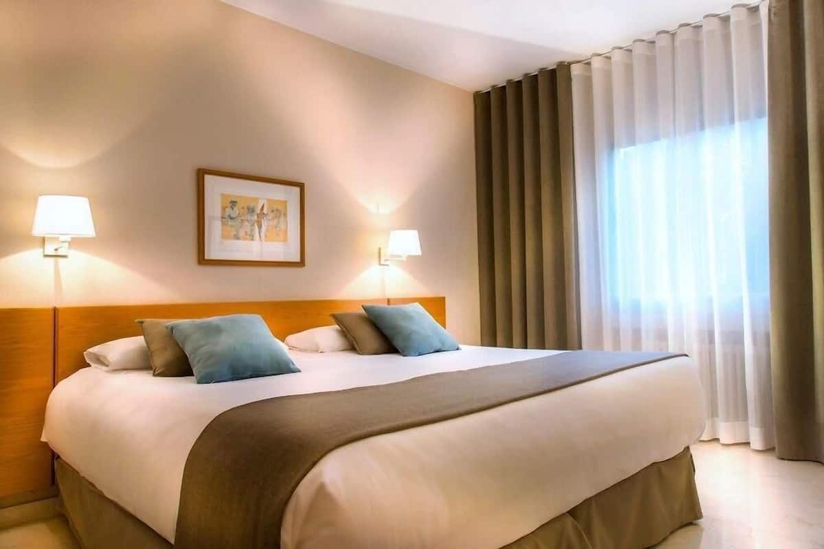 Une chambre d’hôtel avec un grand lit, deux lampes de chevet, un tableau encadré au mur et une fenêtre avec des rideaux.