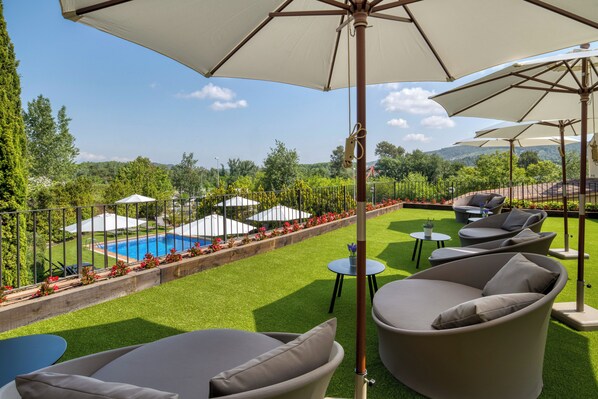 Zona junto a la piscina con mobiliario exterior y sombrillas, rodeada de vegetación y vistas a una piscina.