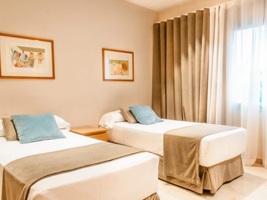 Una habitación de hotel con dos camas individuales, una mesita de noche, un cuadro enmarcado en la pared y una ventana con cortinas.
