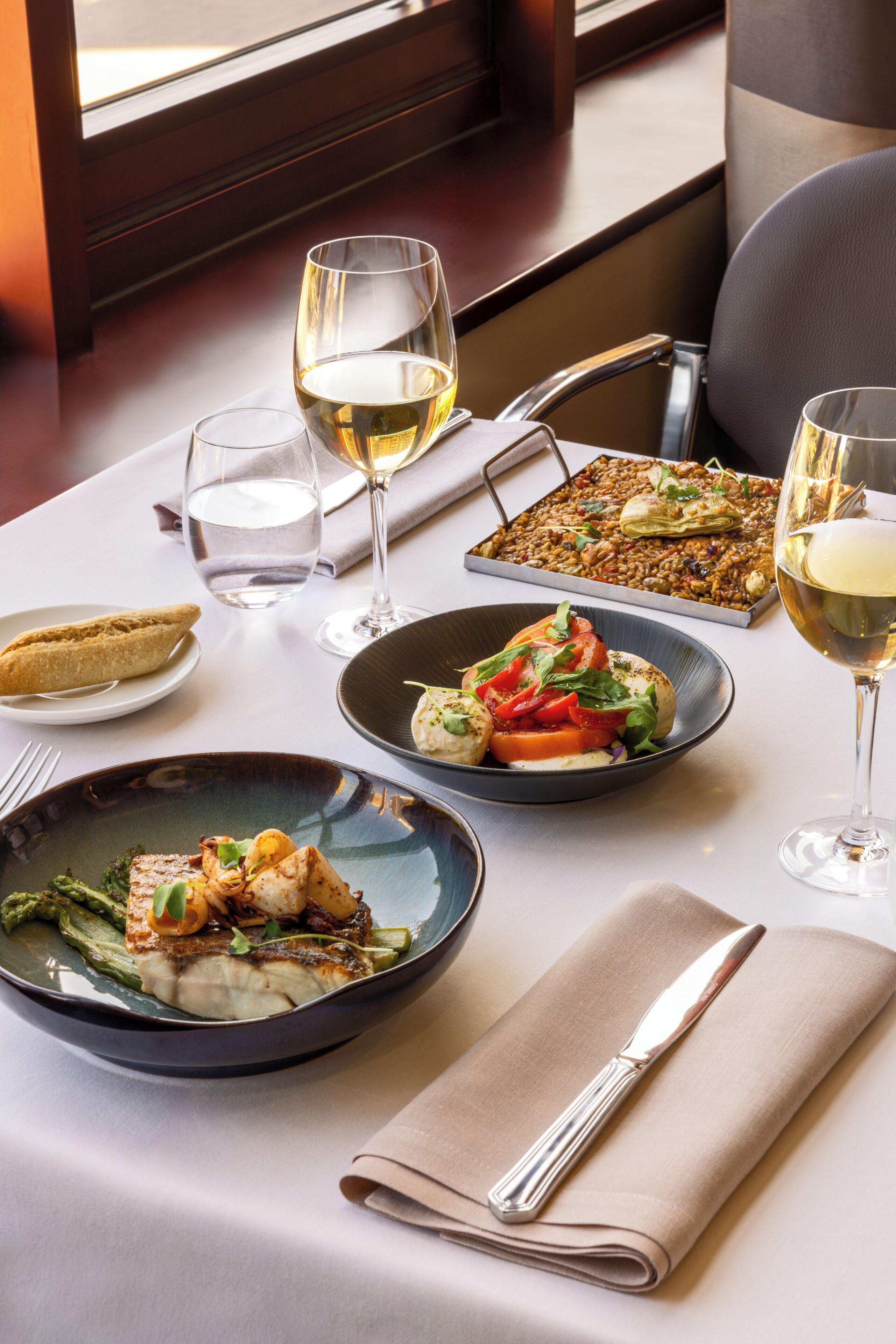 Una mesa de comedor con diversos platos, copas de vino blanco y una servilleta doblada con un tenedor y cuchillo de plata.