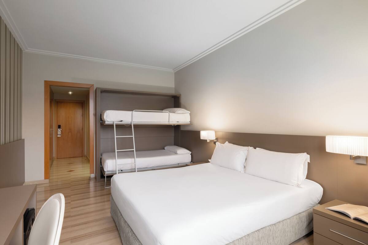 Een hotelkamer met een groot bed, een bureau, een stoel, een stapelbed en een deur naar een andere kamer.