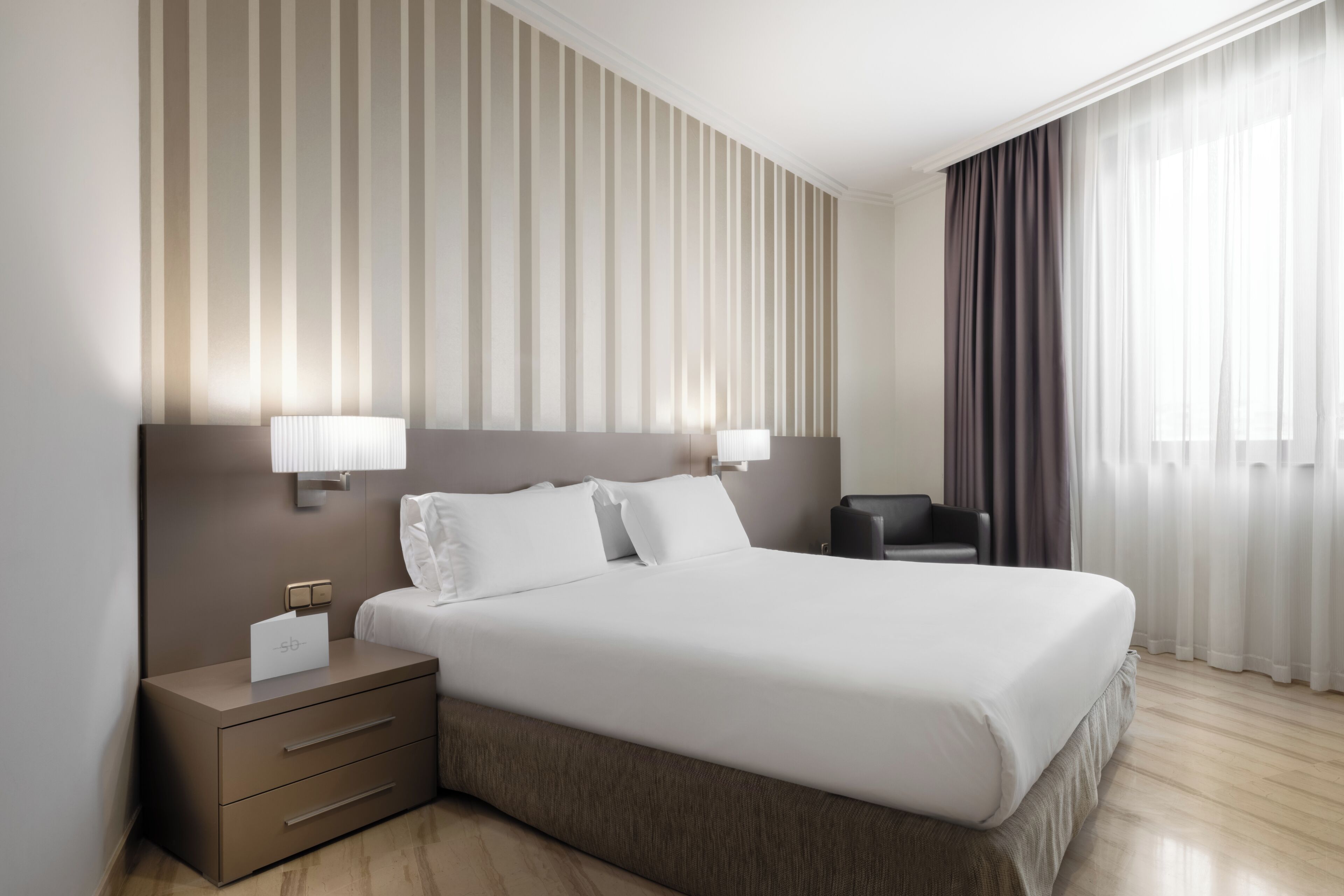 A hotel room with a large bed, bedside lamps, a nightstand, a chair, and a window with curtains.