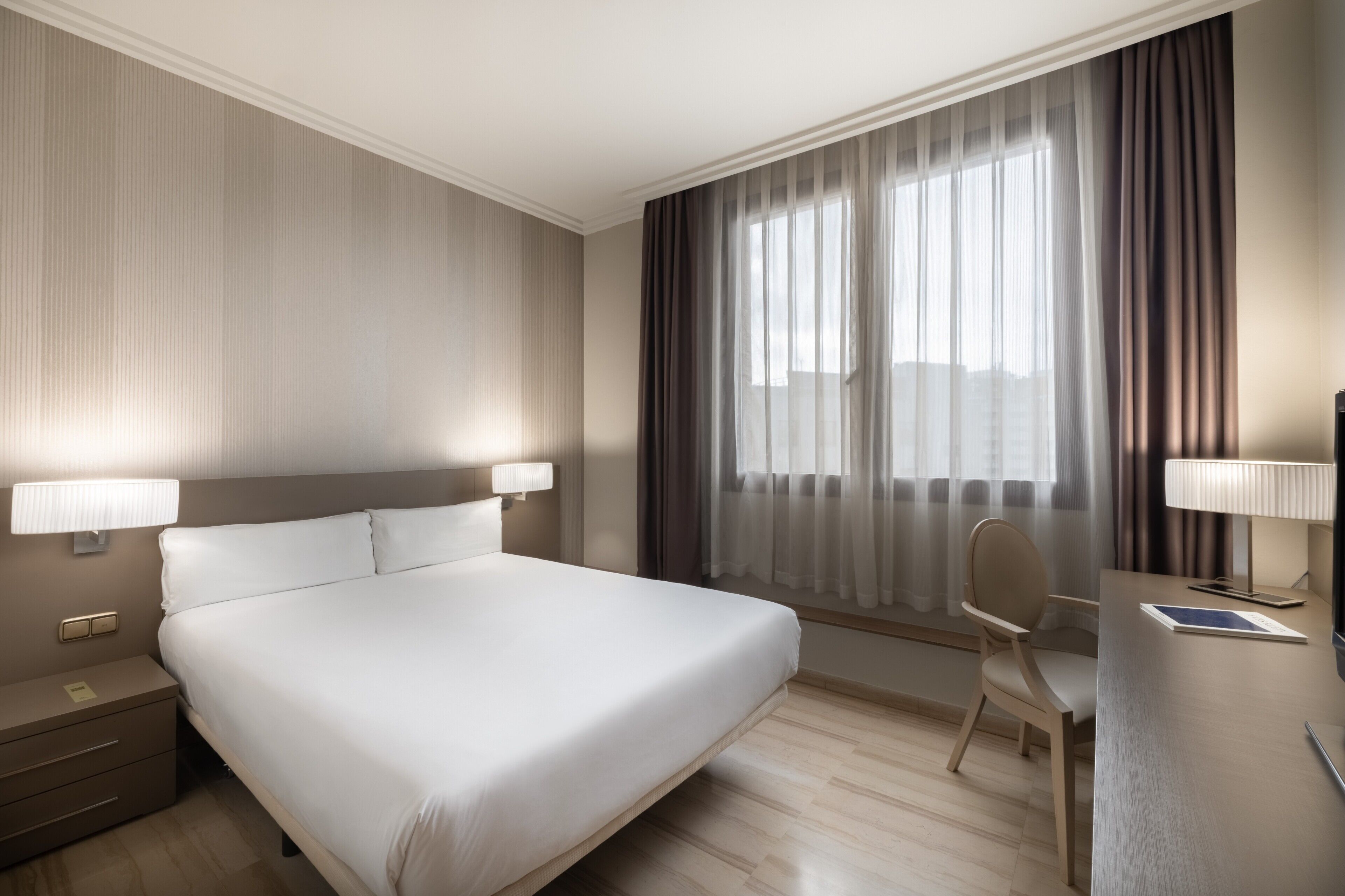Habitación de hotel con una cama grande, un escritorio con silla, una lámpara y un ventanal con cortinas.