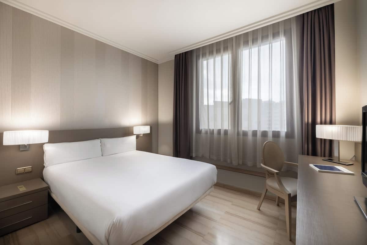 Una habitación de hotel con una cama grande, lámparas de noche, un escritorio con una silla y un ventanal con cortinas.