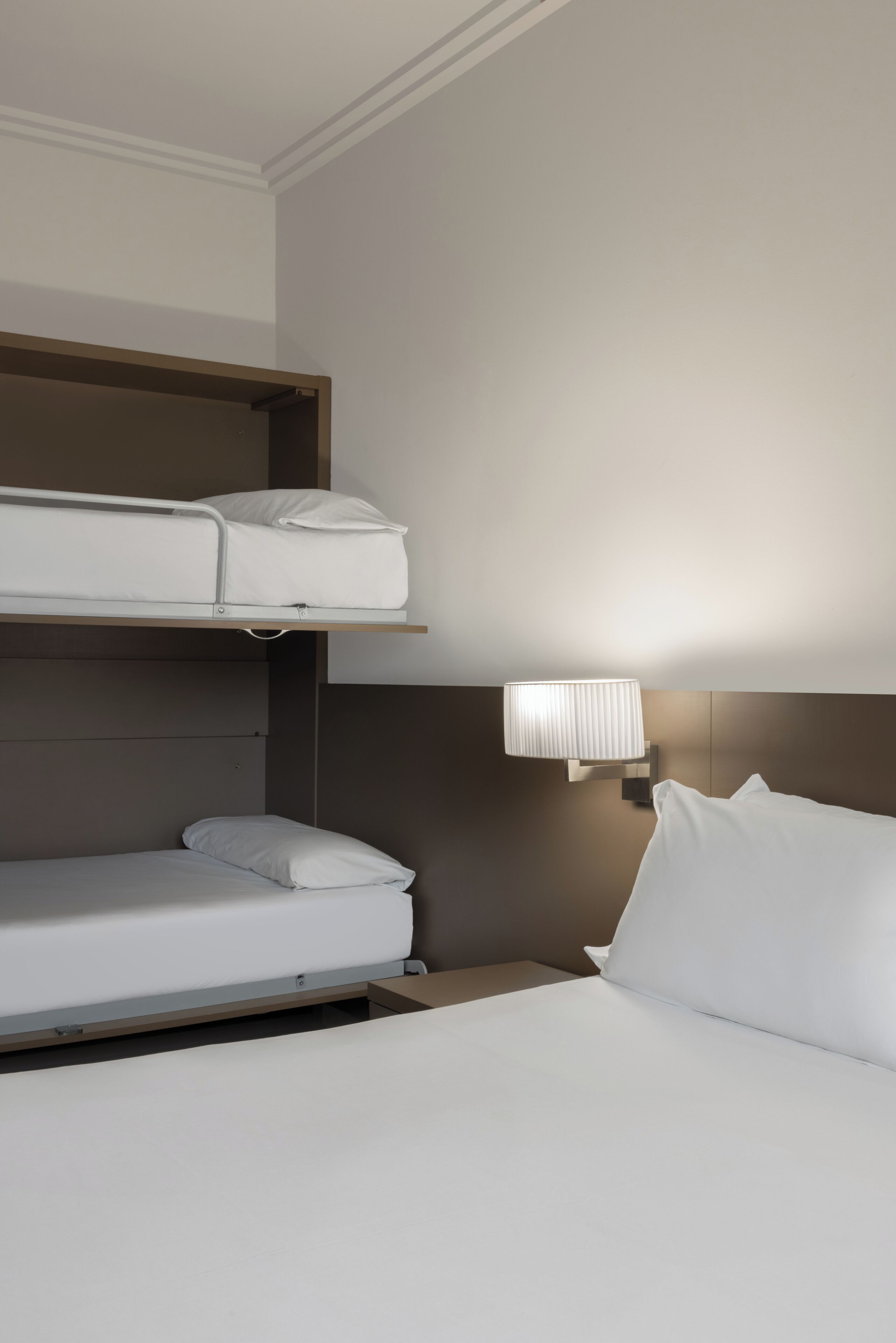 A hotel room with a bunk bed, a single bed, and a wall-mounted lamp.