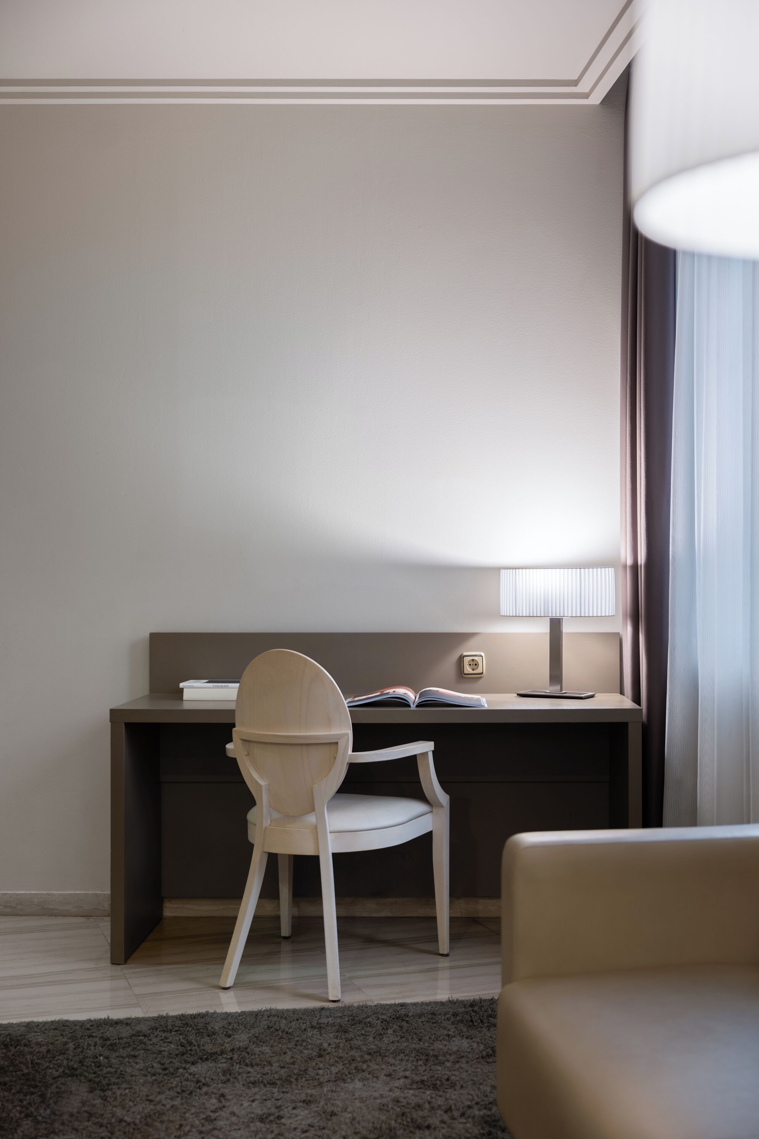 A modern hotel room with a desk, chair, lamp, and a view of the outdoors.