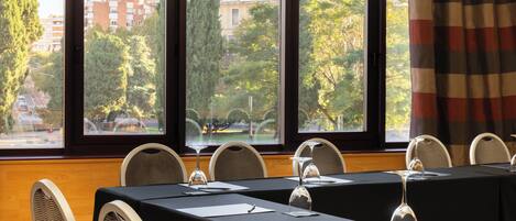 A conference room with a table set for a meeting, large windows with a view of trees and buildings, and a checkered curtain.