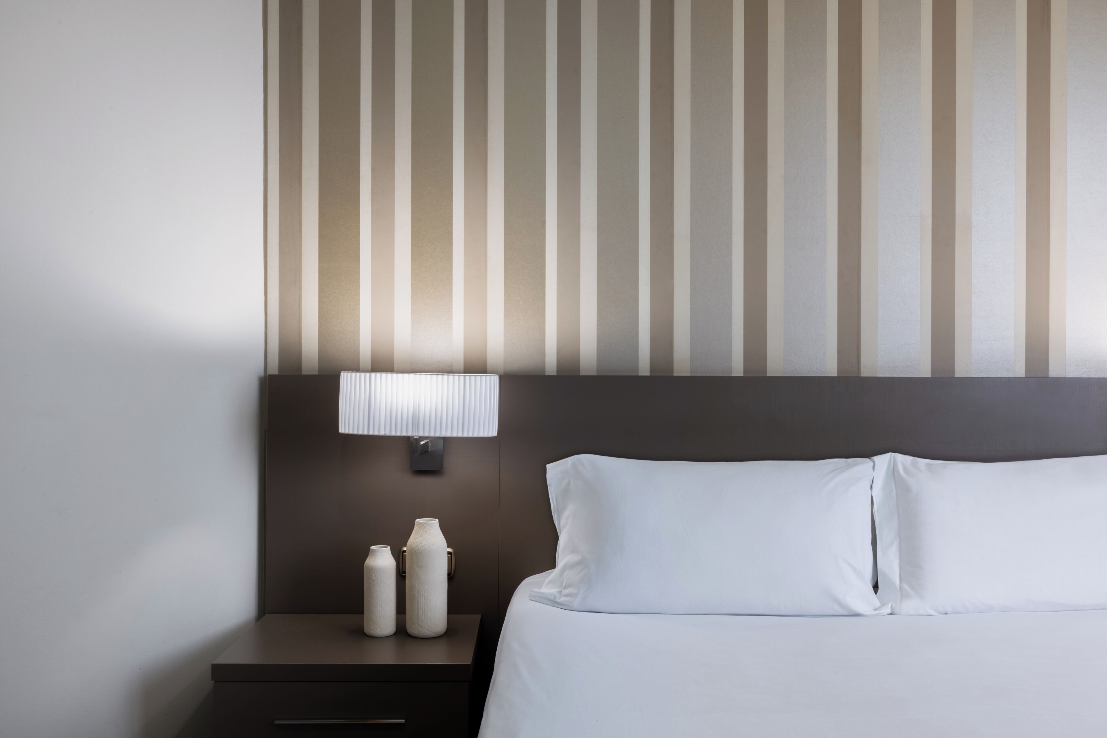 Una habitación de hotel con una cama, una mesita de noche con lámpara y una pared con rayas verticales.