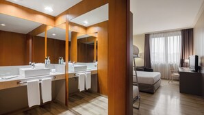 Ein Hotelbad mit einem großen Spiegel, zwei Waschbecken und einem Bett im angrenzenden Zimmer.