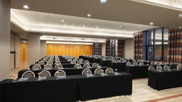 A conference room with rows of tables and chairs, a podium, and a large window with curtains.