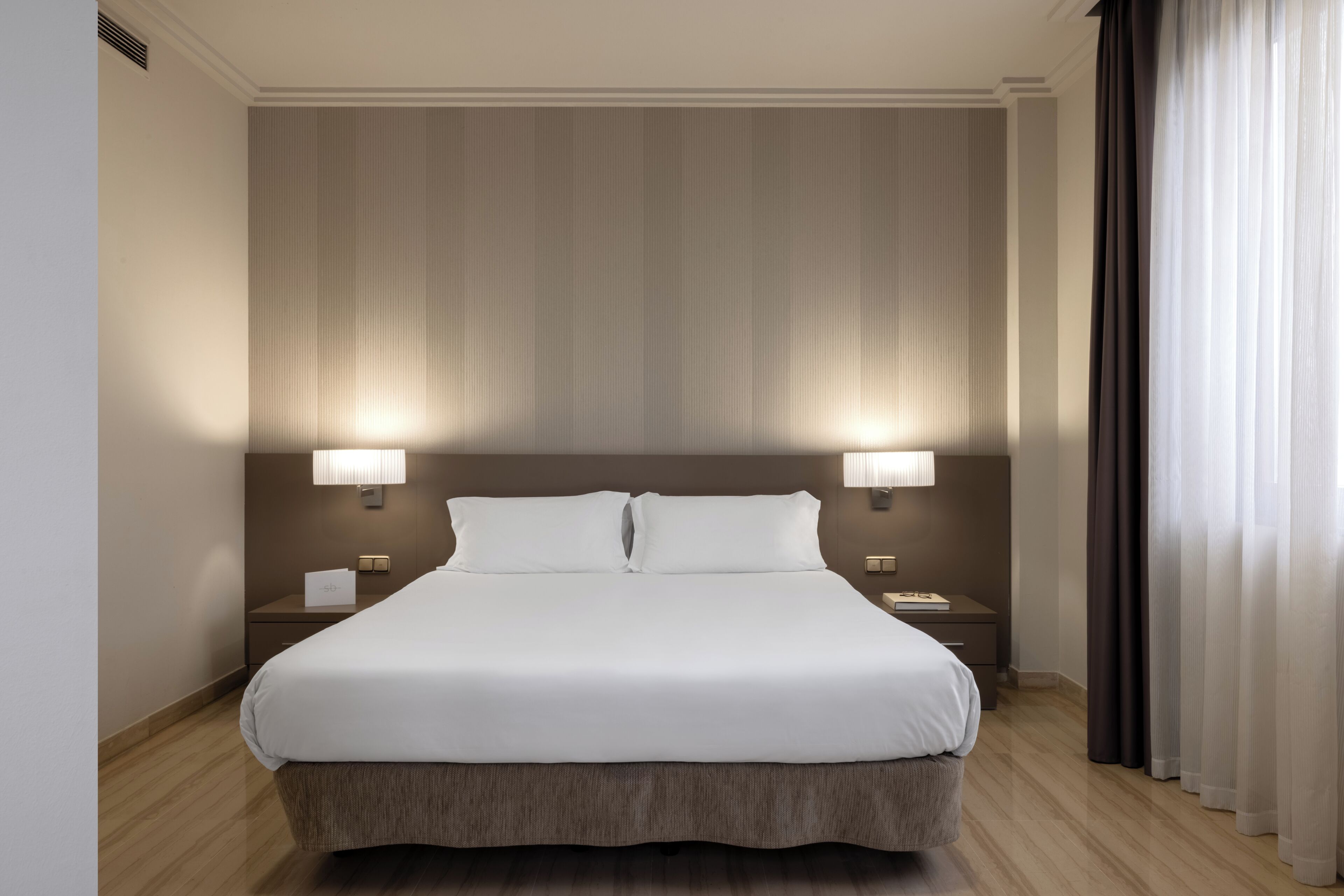 Habitación de hotel con una cama grande, dos lámparas de noche, una mesita de noche y una ventana con cortinas.