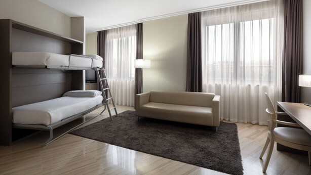 Ein Hotelzimmer mit Stockbett, einer Schlafcouch, einem Esstisch und Blick auf die Stadt durch große Fenster.
