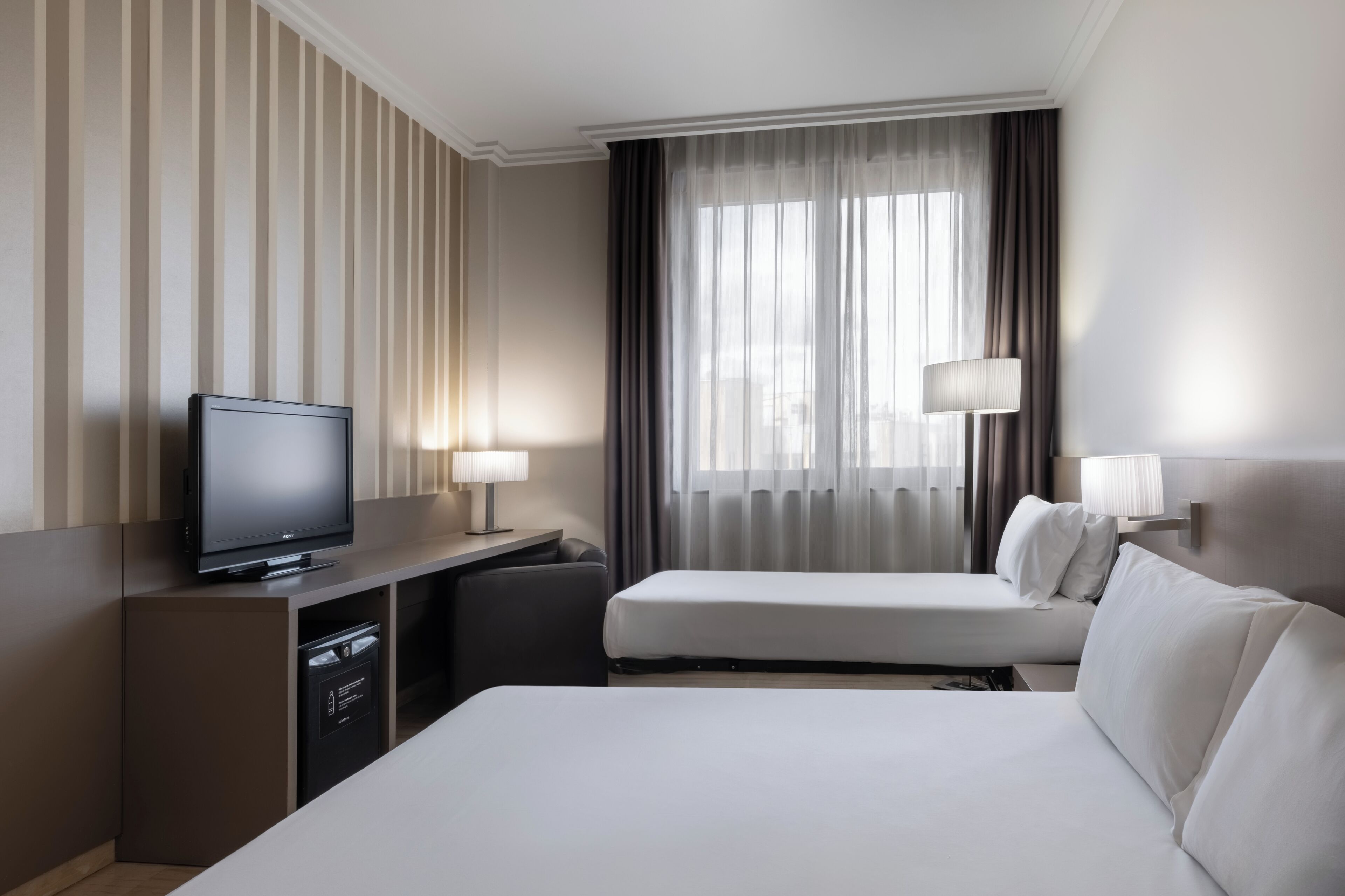 Ein Hotelzimmer mit Flachbildfernseher, Schreibtisch mit Lampe, zwei Betten und einem großen Fenster mit Vorhängen.