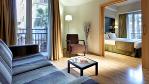 A hotel room with a sofa, armchair, and a small table with a magazine. There is a bed with a green bedspread and a lamp on a side table. - Hotel Dorma Tartessos (Huelva)