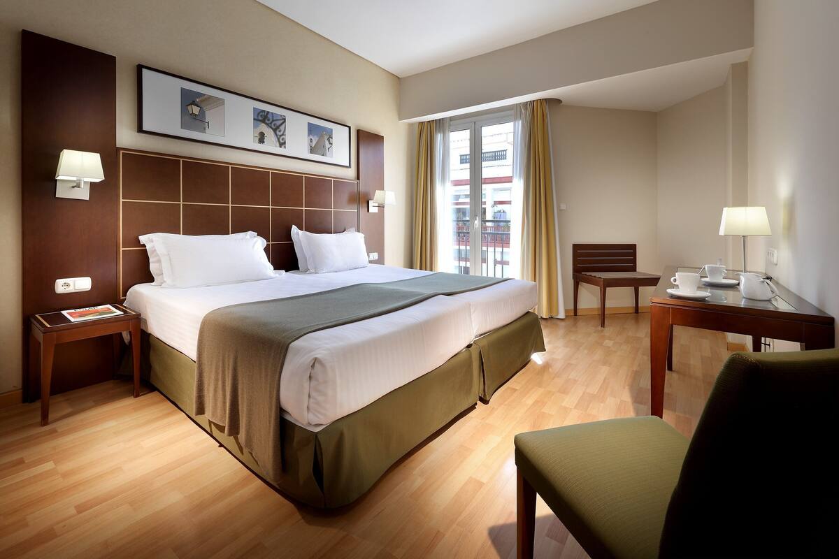 Une chambre d’hôtel avec un grand lit, un bureau avec une chaise, une fenêtre avec des rideaux et une lampe fixée au mur.