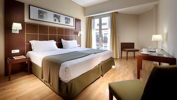 A hotel room with a large bed, a desk with a chair, a window with curtains, and a wall-mounted lamp.