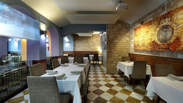 Restaurante com piso xadrez, toalhas de mesa brancas e um mural de tijolos na parede.
