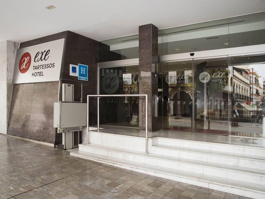 The exterior of the Excé TARTESSOS HOTEL with a modern facade, large glass entrance, and a visible staircase leading to the entrance.
