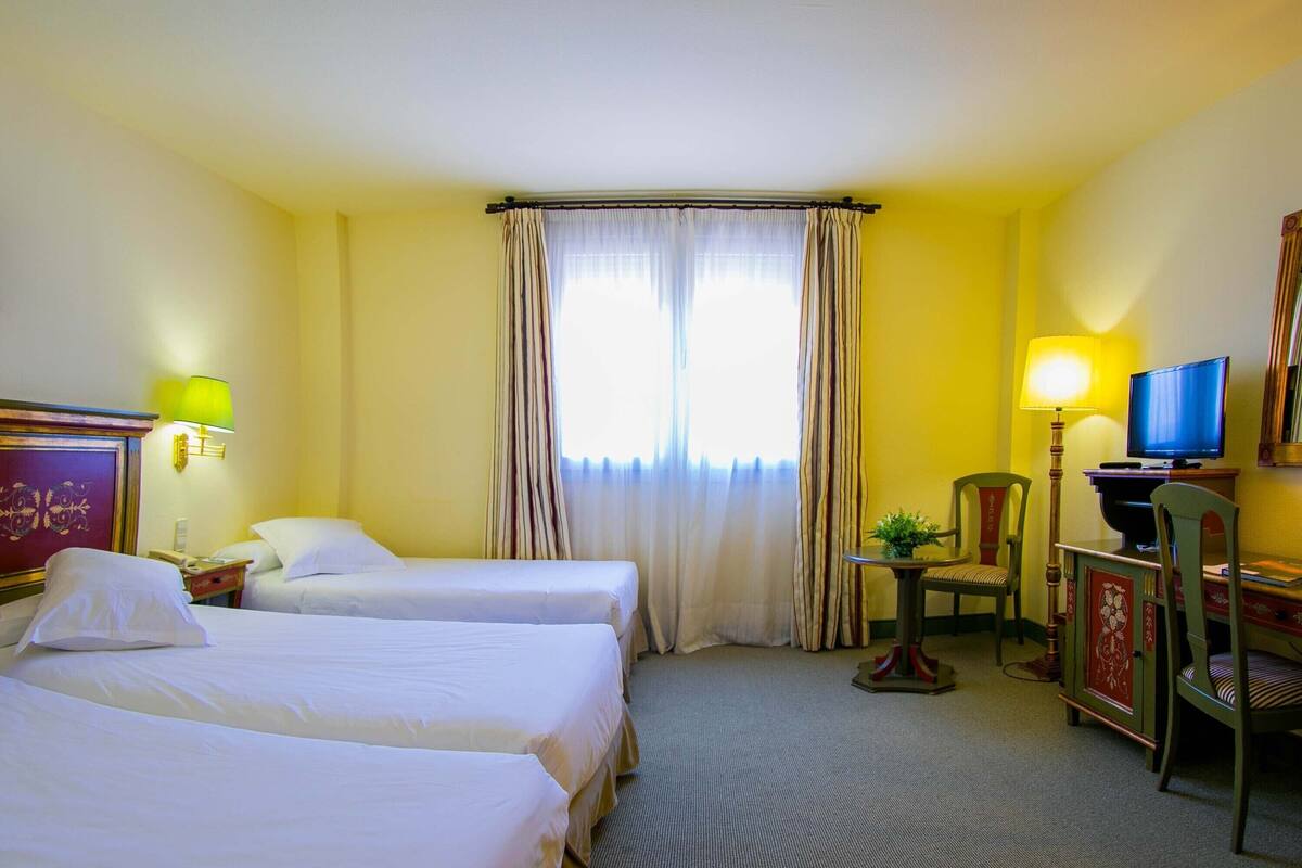 Quarto de hotel com duas camas, uma mesa com cadeira, televisão e janela com cortinas.