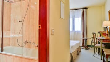 Habitación de hotel con ducha de vidrio, cama, una mesita con lámpara y una silla.