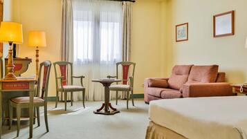 Habitación de hotel con una cama, dos sillones, una mesita, un escritorio con lámpara y una ventana con cortinas.
