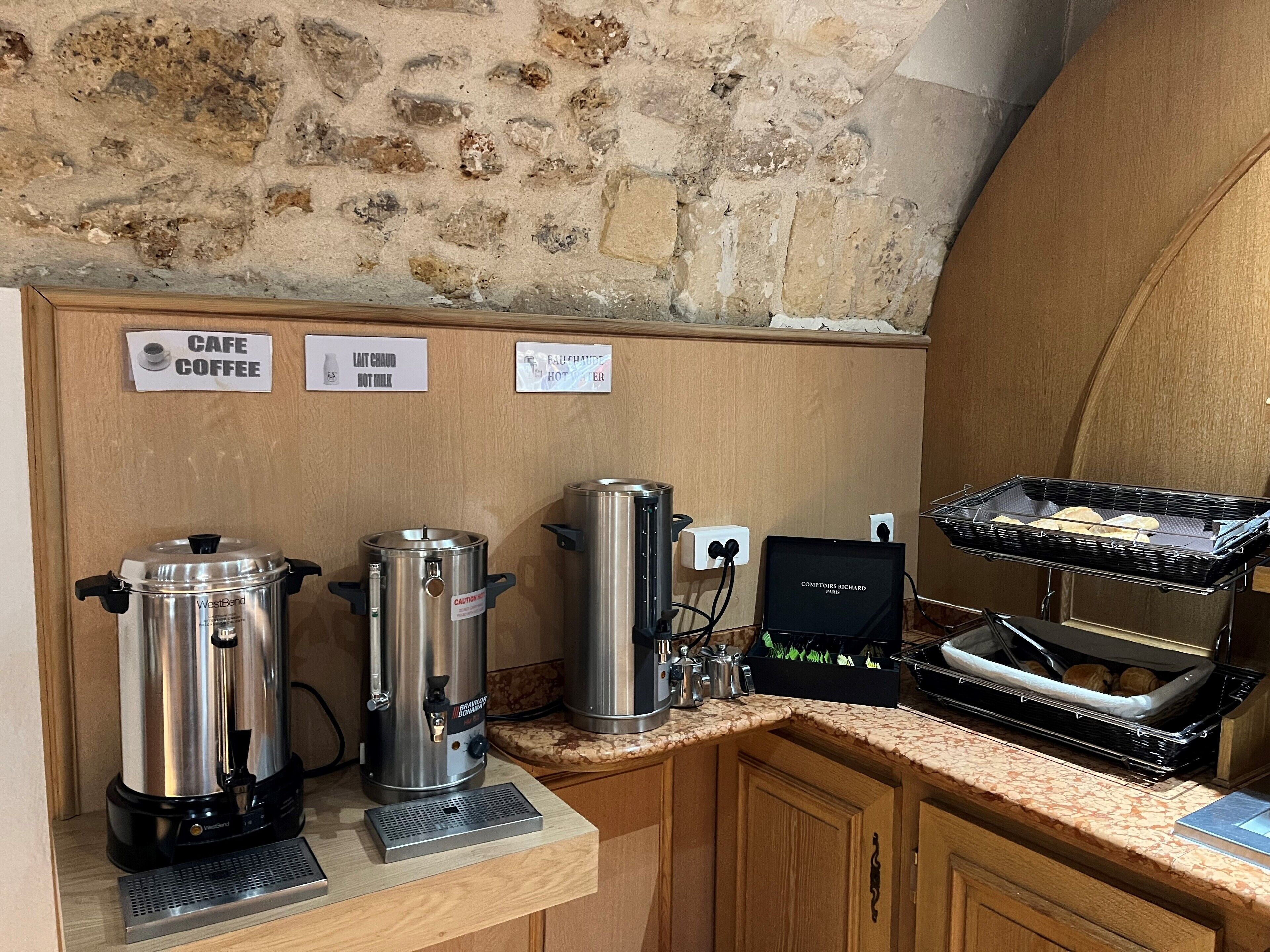 Una piccola cucina accessoriata con una macchina del caffè, un distributore di latte caldo e un tostapane. Si notano cartelli con scritto 'Cafe Coffee' e 'Lait chaud Hot Milk'.