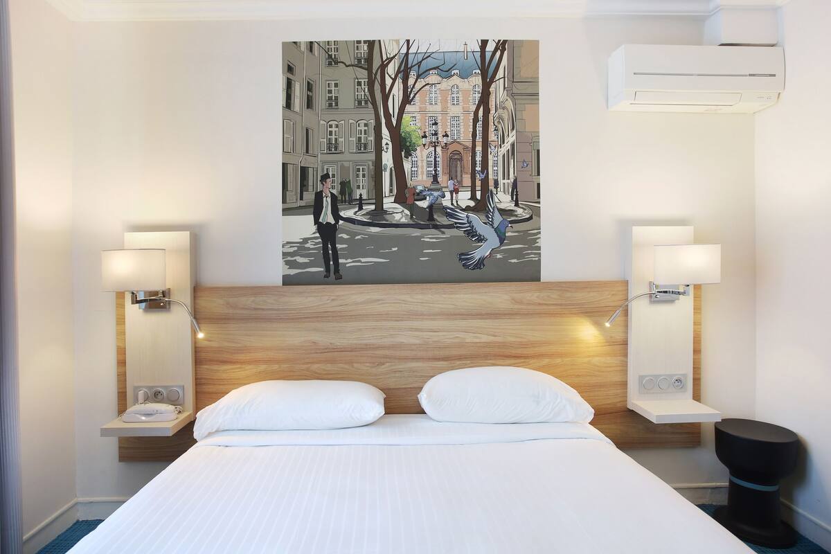 A hotel room with a wooden headboard, a bed with white linens, two bedside tables with lamps, a wall-mounted air conditioner, and a cityscape painting above the bed.