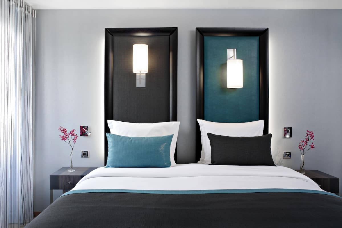 Une chambre d’hôtel moderne avec un grand lit, deux tables de chevet, une petite table, une lampe fixée au mur et une fenêtre avec des rideaux.