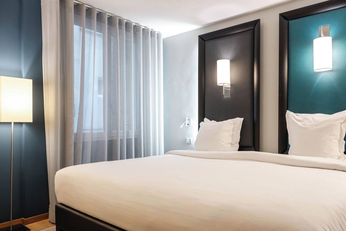 Une chambre d’hôtel avec un grand lit, une table de chevet, une lampe, une fenêtre avec des rideaux et un luminaire fixé au mur.