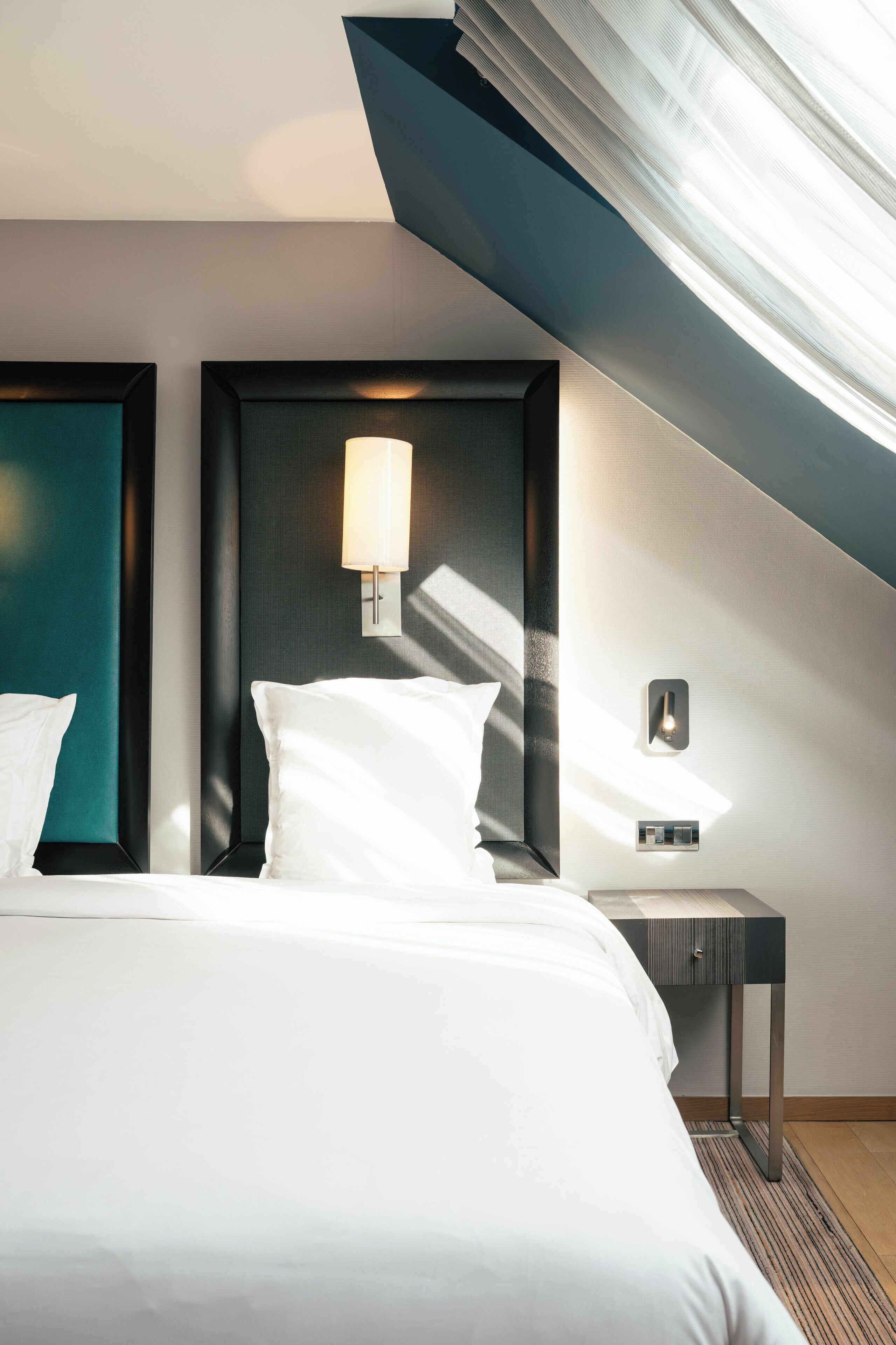 Habitación de hotel con una cama, una mesita de noche, una lámpara de pared y vistas del cielo a través de una ventana.