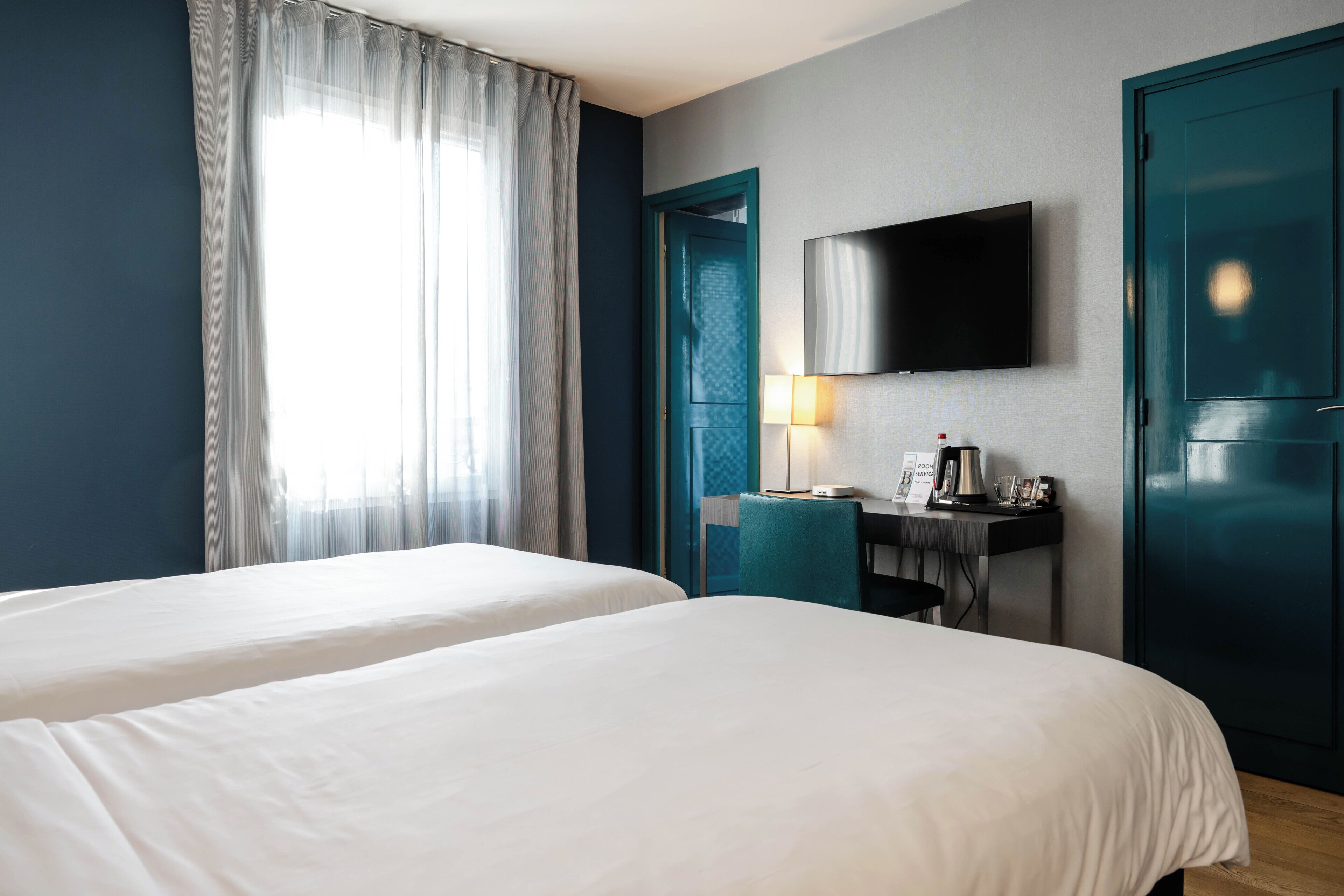 A hotel room with two beds, a desk with a lamp, a flat-screen TV, and a door with a teal frame.