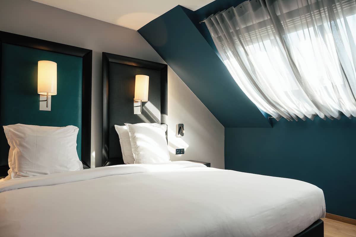 Une chambre d’hôtel avec un lit, deux lampes fixées au mur, une fenêtre avec des rideaux légers et un mur d’accent de couleur turquoise.