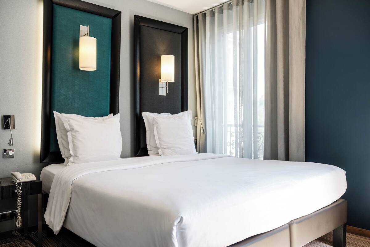 Une chambre d’hôtel avec un grand lit, deux lampes de chevet, un téléphone et une vue sur l’extérieur à travers des rideaux légers.