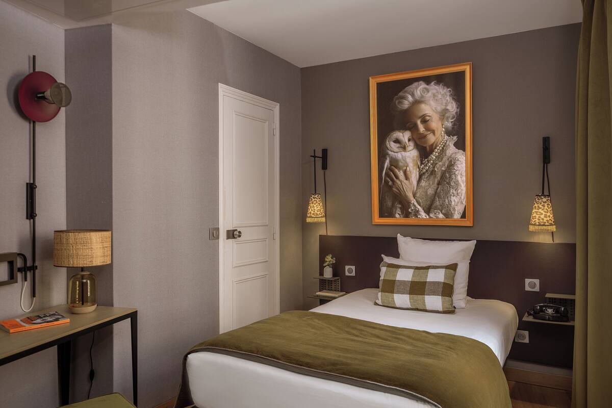 A hotel room with a bed, bedside tables, a desk, a lamp, a framed picture on the wall, and a door.