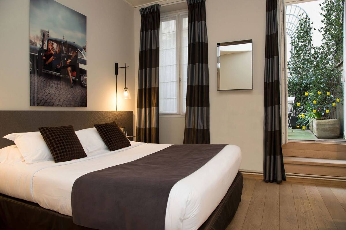 A hotel room with a large bed, a nightstand, a wall-mounted lamp, a window with curtains, a mirror, and a view of greenery outside.