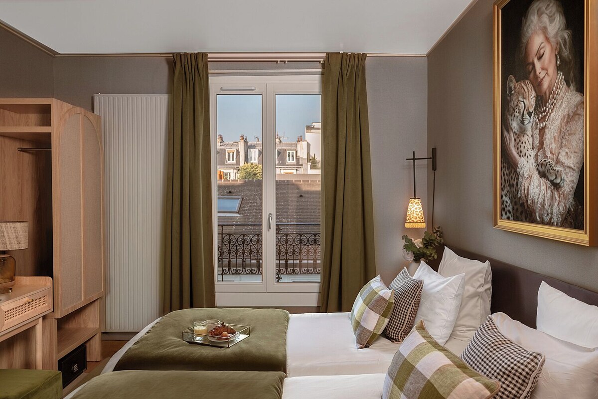 A hotel room with two beds, a wooden wardrobe, a desk with a lamp, a painting, and a view through a window with curtains.