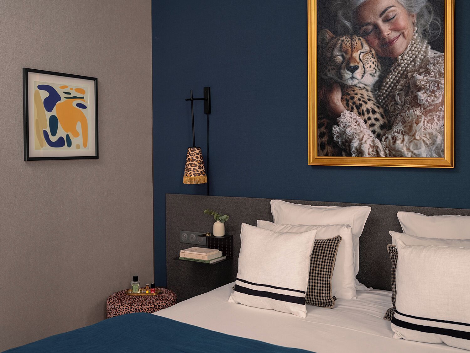 A modern bedroom with a large bed, a framed abstract painting, a leopard-print lamp, and a framed photograph of a woman with a cheetah.