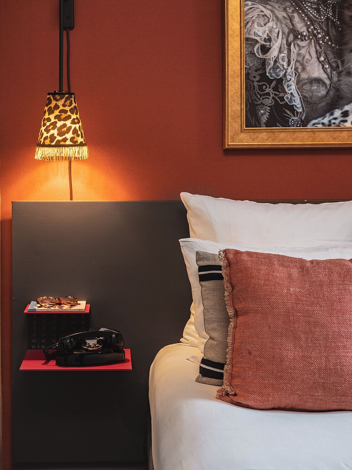 A neatly made bed with white and pink pillows, a lamp with a leopard print shade, and a framed picture on the wall.