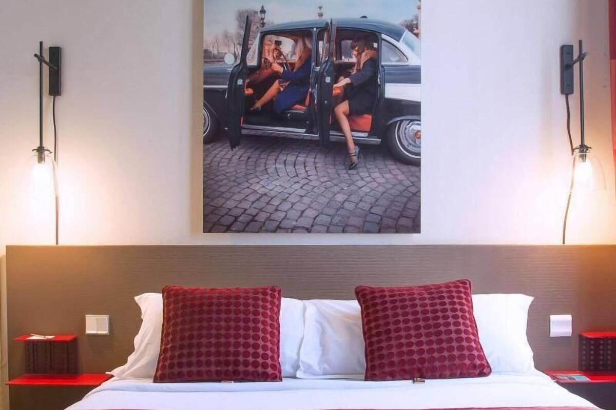 A hotel room with a large bed, two red pillows, a red blanket, a nightstand with a lamp, and a wall-mounted picture of a vintage car.