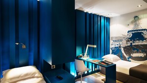 Una habitación de hotel moderna con una decoración azul, un escritorio con lámpara, una silla y dos camas.