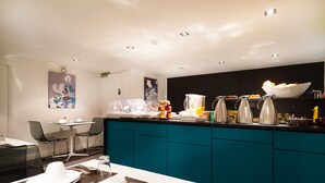 A modern kitchen with teal cabinets, a dining area with a table set for two, and a coffee station with multiple coffee makers. - Hotel Arc de Triomphe Etoile (Paris)