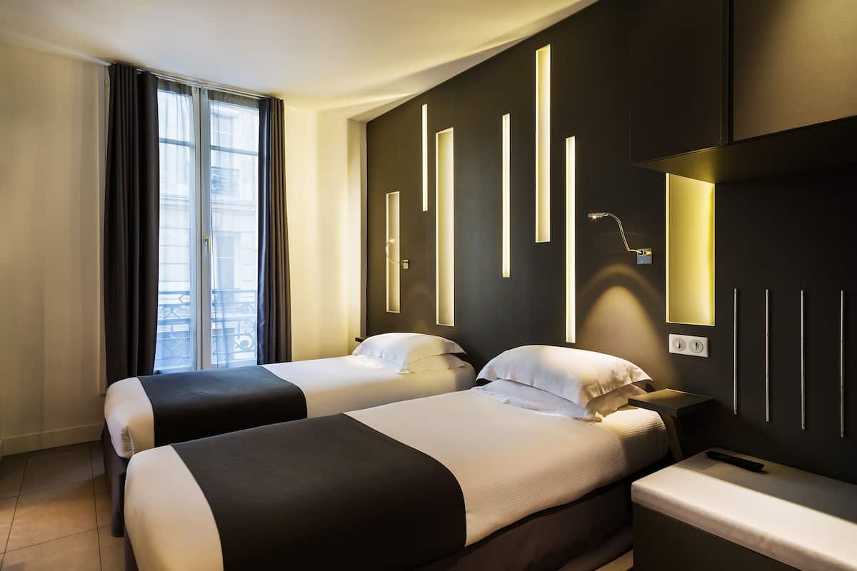 Habitación de hotel con dos camas, una mesita de noche, televisor montado en la pared y ventana con cortinas.