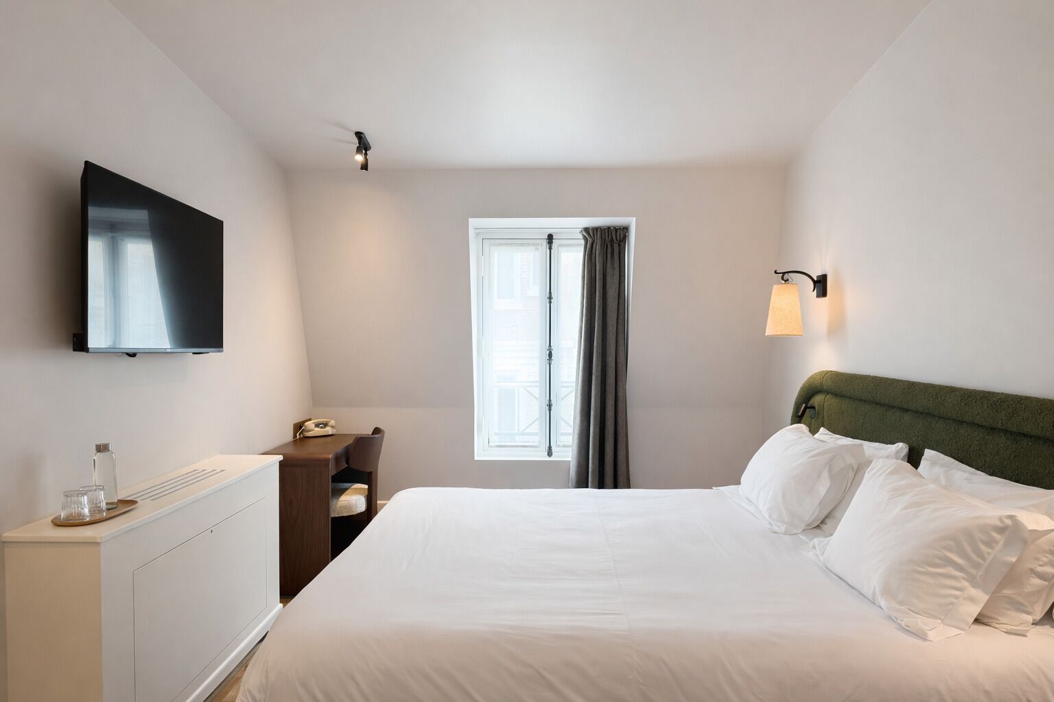 A hotel room with a bed, a desk, a chair, a TV, a window with curtains, and a wall-mounted light.